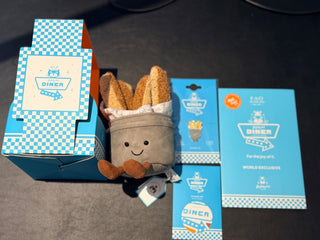 A plush toy dressed as a french fries character in a gray bucket, displayed with its blue and white checkered branded packaging on a dark surface.