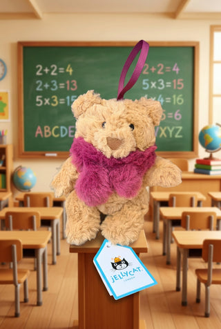 A small plush teddy bear ornament in tan with a bright pink scarf and hanging ribbon, shown on a wooden surface with a blue container in the background and a Jellycat tag in front.